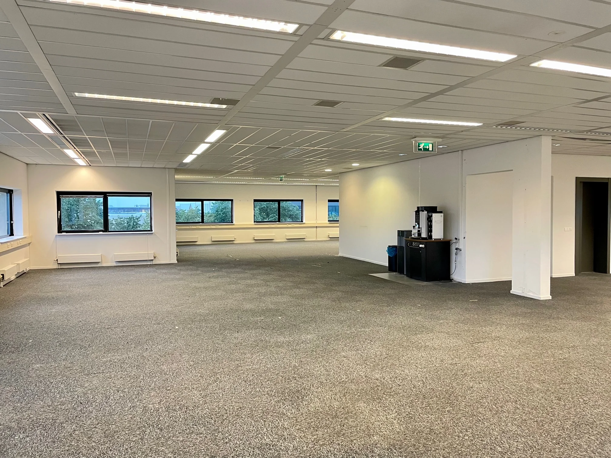 Leeg kantoorpand aan de Karel Doormanweg met tapijt, systeemplafond en een koffieautomaat in de hoek.